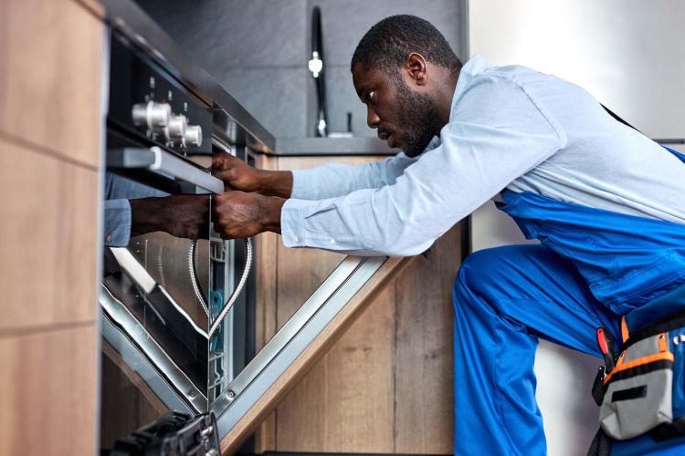Professional Afro Handyman Repairing Dishwasher, Need To Change Old Dishwasher Hose Professional Afro Handyman Or Contractor Repairing Dishwasher, Need To Change Old Dishwasher Hose, Black Guy In Blue Overalls Is Concentrated On Work, In Kitchen Indoors. Side View Portrait.
